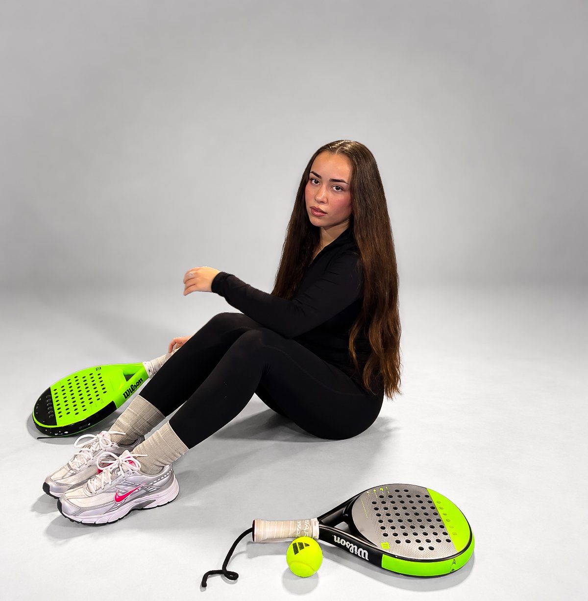 Ceyda with long brown hair in a black sports outfit, sitting on the floor next to two padel rackets and a ball against a light studio backdrop